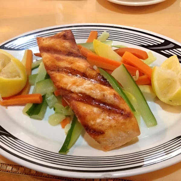 Grilled salmon fillet with mixed vegetables and lemon wedges on a white plate with black stripes.